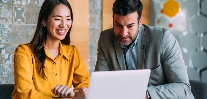 A businesswoman and businessman sitting at a table smiling while looking at a laptop