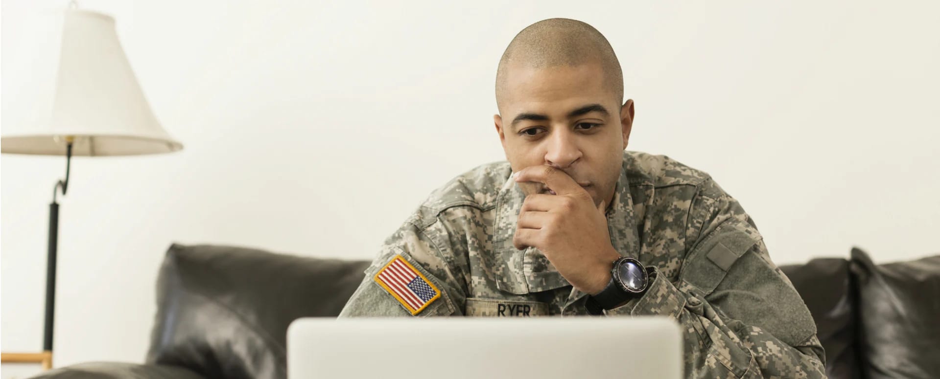 Male in military uniform sitting on a black leather couch with a laptop in front of him.
