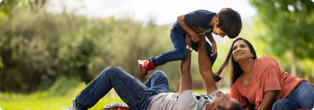 A man, laying on a blanket on grass, holding a young boy over his head, and a woman lies beside the man smiling