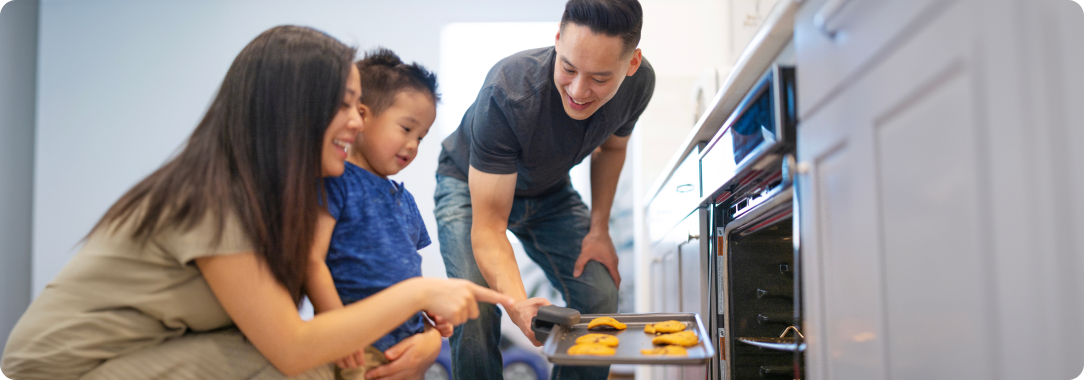 A woman, bending down next to a young child, pointing at food on a cooking sheet that a man is taking out of an oven