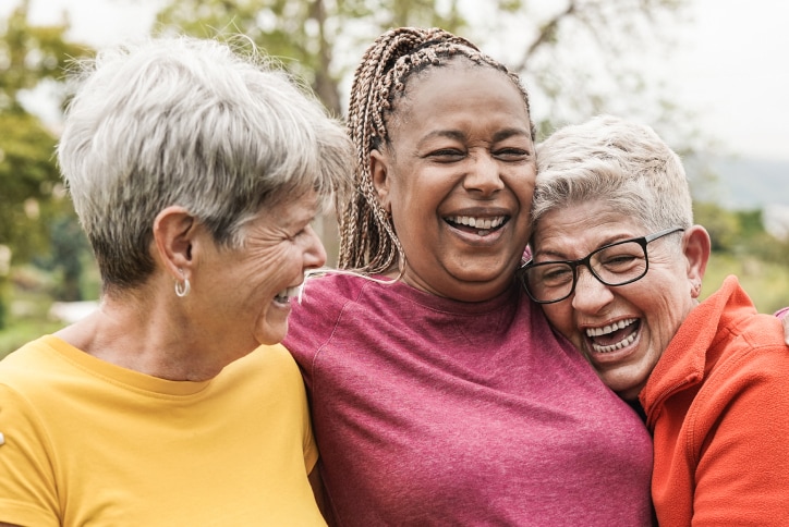 three older woman hugging and laughing together while wearing a yellow, pink and orange top