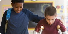 A boy helping another boy while walking on crutches in a classroom 