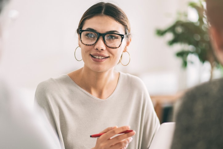 Young woman holding red pen in hand with glasses on speaking to two other individuals across from her.