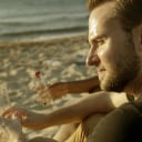 A young man sitting on a beach overlooking the ocean with a bonfire in front of him and two friends on either side of him