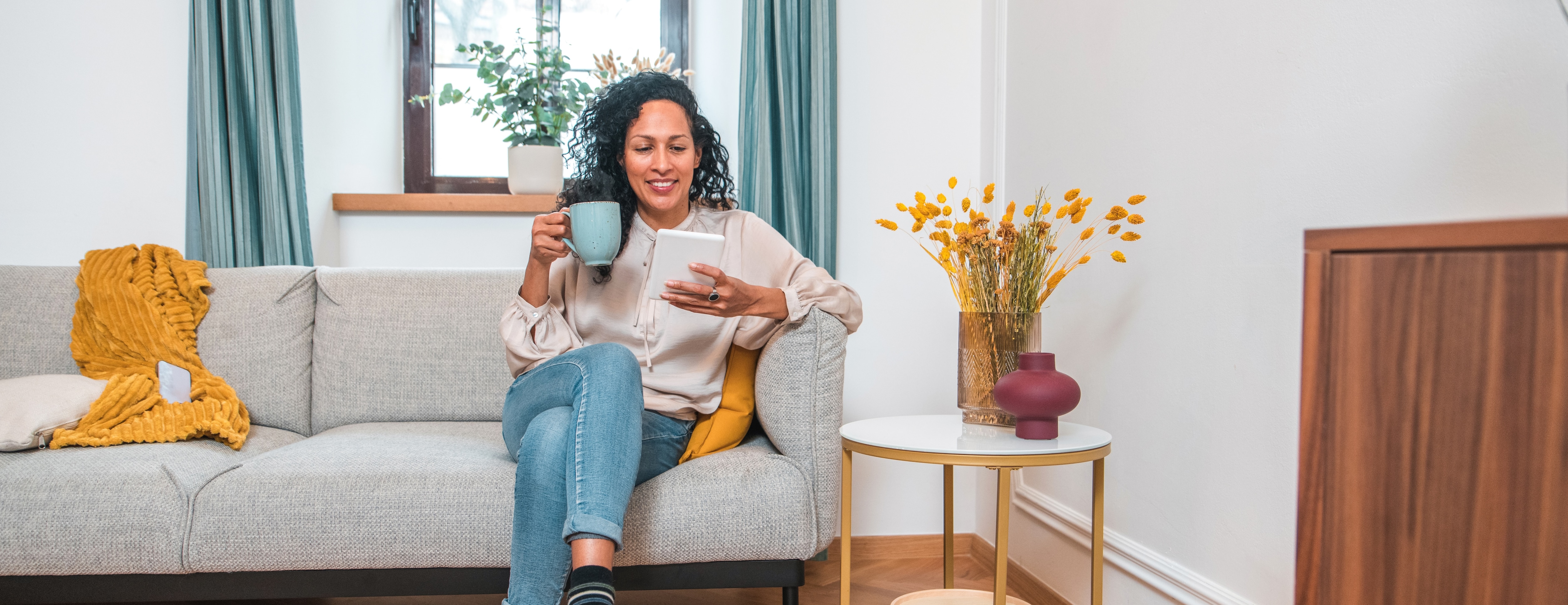 Woman in jeans and long sleeve top holding a blue mug while sitting on a gray couch.