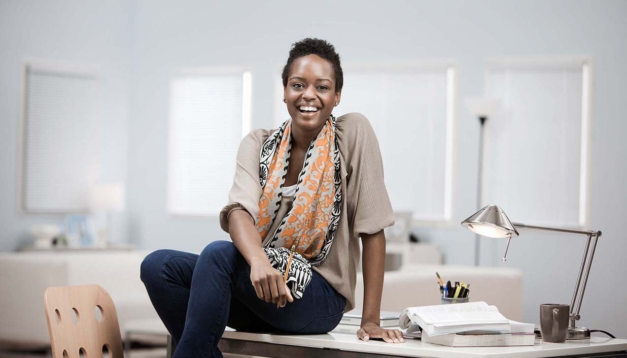 woman sitting on home desk wearing a light brown short sleeve top with a colorful scarf while smiling at camera