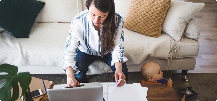 Woman sitting on a white couch typing information onto a laptop while a baby sits on the floor next to her