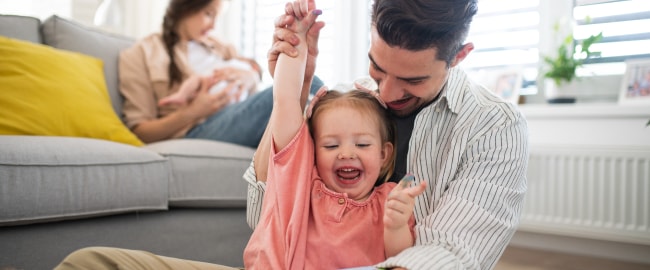 A man raising a little girl's arm while she sits in his lap laughing. A woman sits on a couch behind them holding a baby.