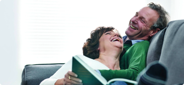 A woman, reading a book, laying against a man, looking up at the man, both of them laughing