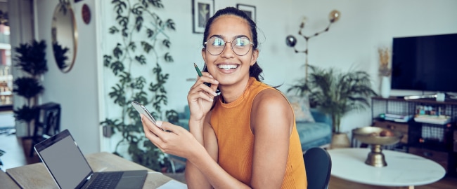 A woman sitting in front of a laptop, holding a cellphone, smiling