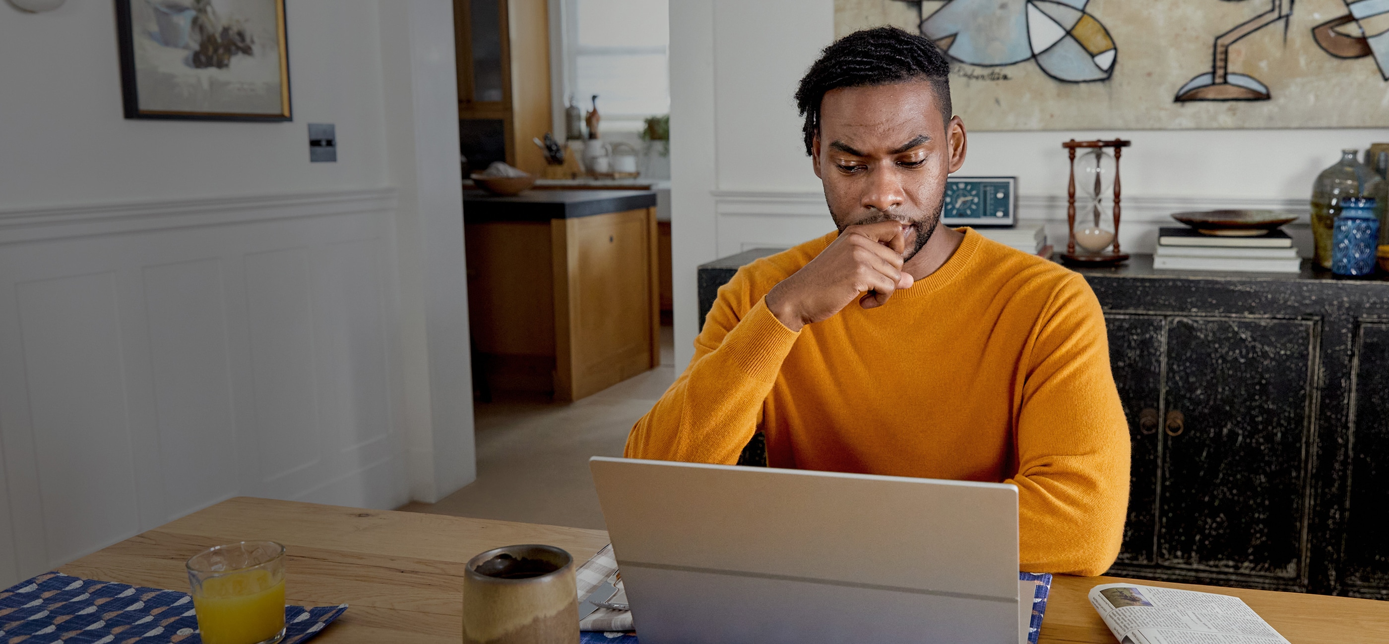 male sitting at wooden table in long sleeve orange sweater staring at laptop screen