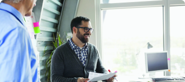 A man wearing glasses and a gray cardigan sweater smiling at another person in front of him, holding a stack of paper