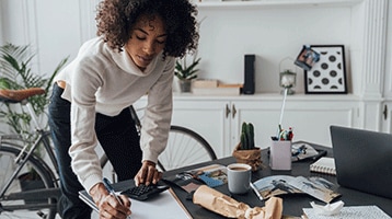 Young African American woman leaning over a home office desk writing on a piece of paper.