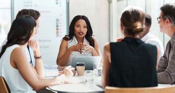 six staff members sitting at round table focused on female speaker in white top with black dots