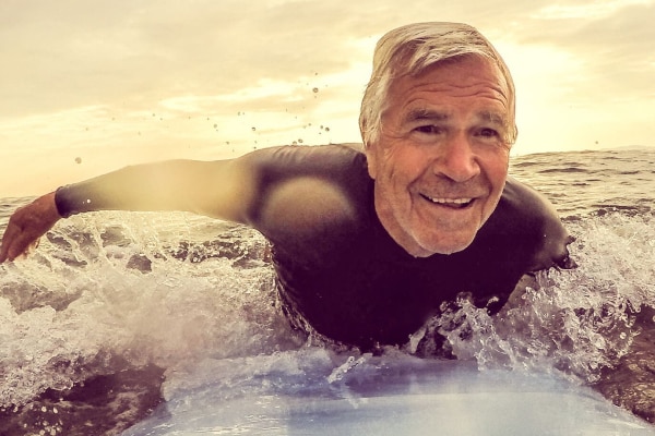 An older man smiling, laying on a surfboard, paddling through water