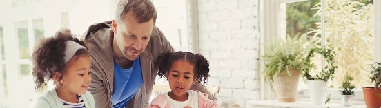 A middle-aged man in the middle of two young girls while they all look down at a book, in a kitchen