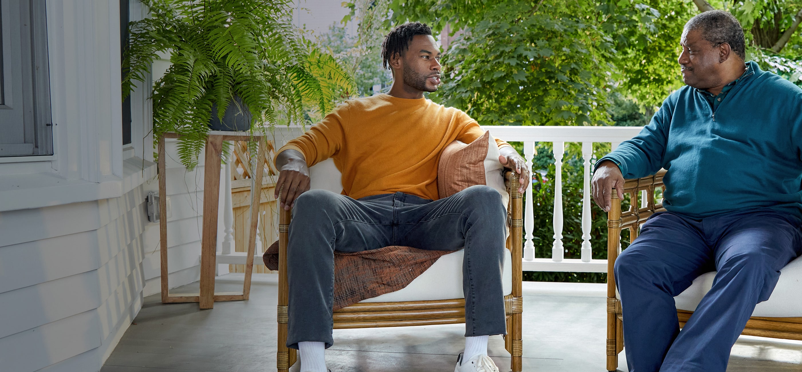 male in orange long sleeve sweater sitting in chair on porch talking to older male in blue button up long sleeve