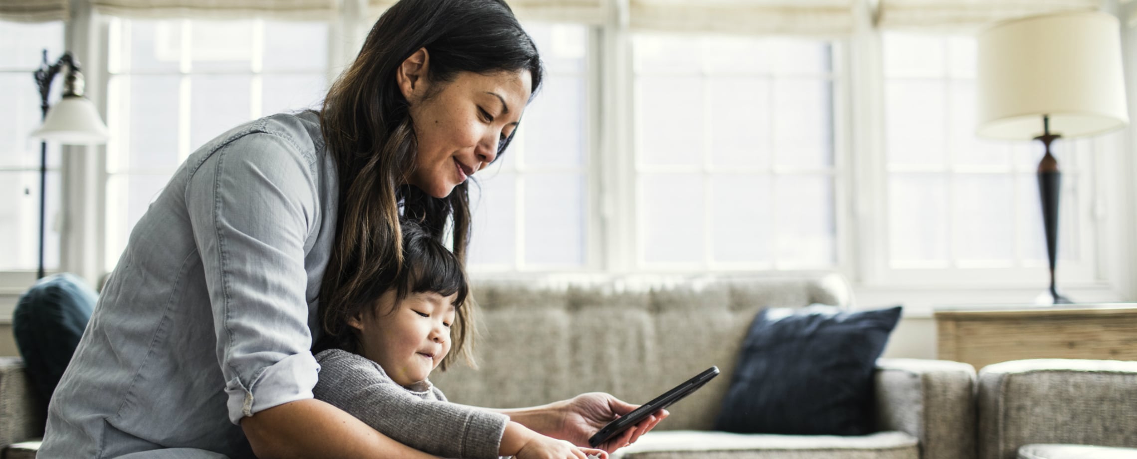 Woman sitting with child standing between her arms while they look at a cell phone