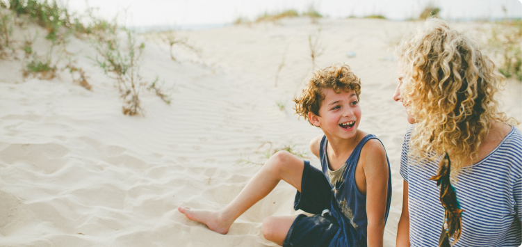 Mother and son smiling at each other while sitting on sand at a beach